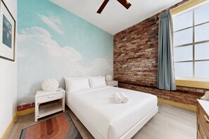 A hotel room with a brick wall, a bed with white linens, a nightstand, a small table, and a window with curtains. - Serenity Cloud Apartment in Vibrant Footscray (Melbourne)