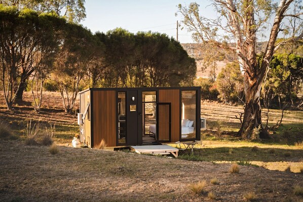 A small, modern cabin with a wooden exterior and glass doors, situated in a natural landscape with trees and open fields.