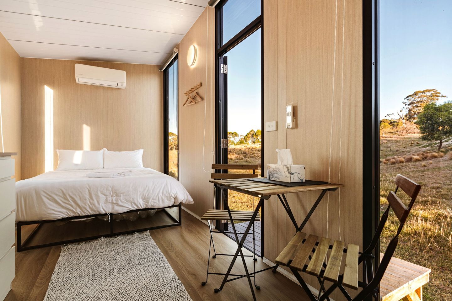 A bedroom with a bed, a small table, and a chair, a large window, and a view of the outdoors.