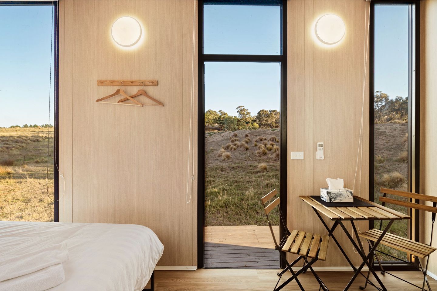 Une chambre à coucher avec un lit, une table et une chaise, et une vue sur un paysage verdoyant par de grandes fenêtres.