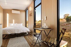A bedroom with a bed, a small table, and a chair, a large window, and a view of the outdoors.