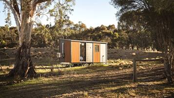 A small, single-story cabin with a wooden exterior, a sliding glass door, and a window, situated in a grassy field with trees in the background.