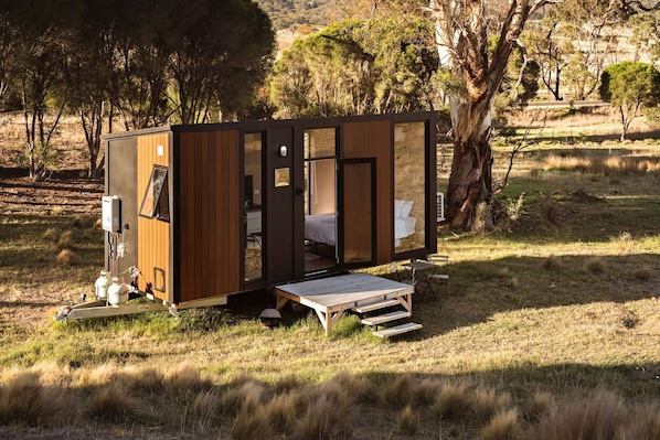 A small, modern cabin with a wooden exterior, a bed inside, and a small deck area.
