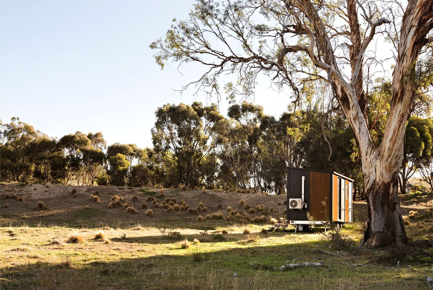 A small cabin with a sloped roof, positioned on a grassy hill with trees in the background.