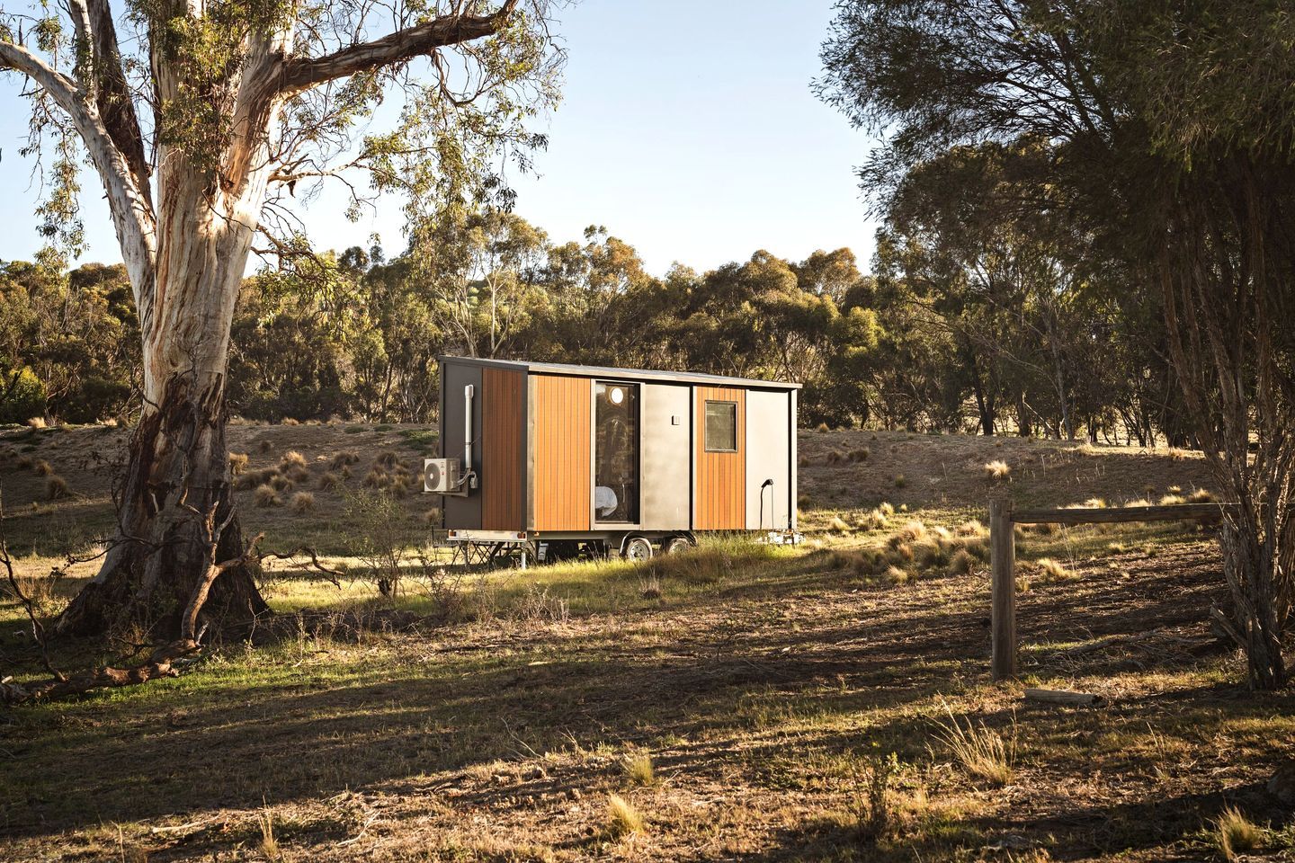 Une petite cabane à un seul étage, avec une façade en bois, une porte-fenêtre coulissante et une fenêtre, située dans un champ herbeux, avec des arbres en arrière-plan.