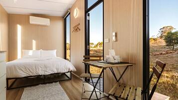 A bedroom with a bed, a small table, and a chair, a large window, and a view of the outdoors.