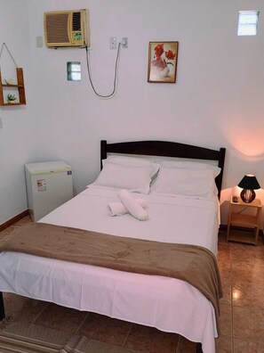 A neatly made bed with white linens and a brown throw blanket, a small bedside table with a lamp, a wall-mounted air conditioner, and a framed picture.