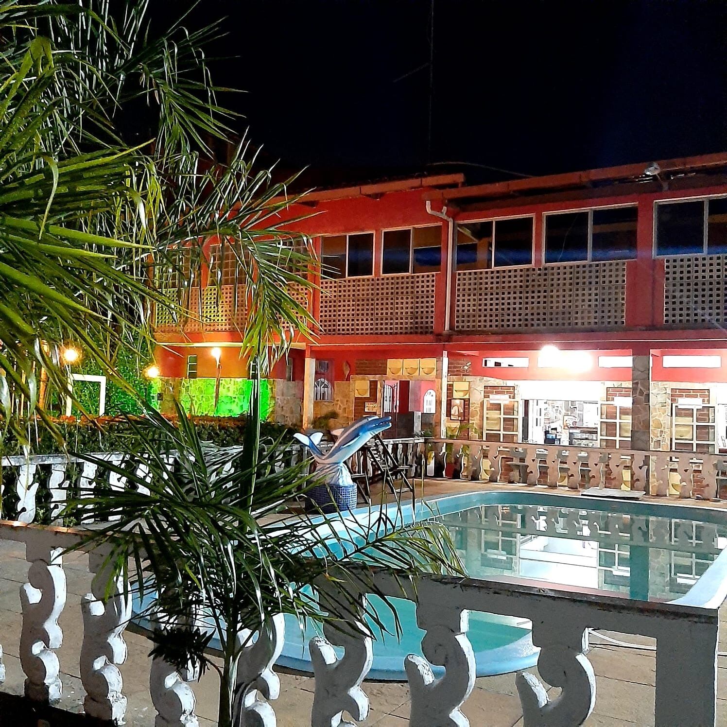 Vista noturna de um edifício com área de piscina, apresentando uma palmeira e um portão decorativo.