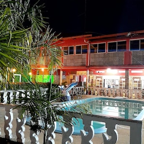A nighttime view of a building with a pool area, featuring a palm tree and a decorative fence. - Pousada Canto de Guarajuba (Camacari)