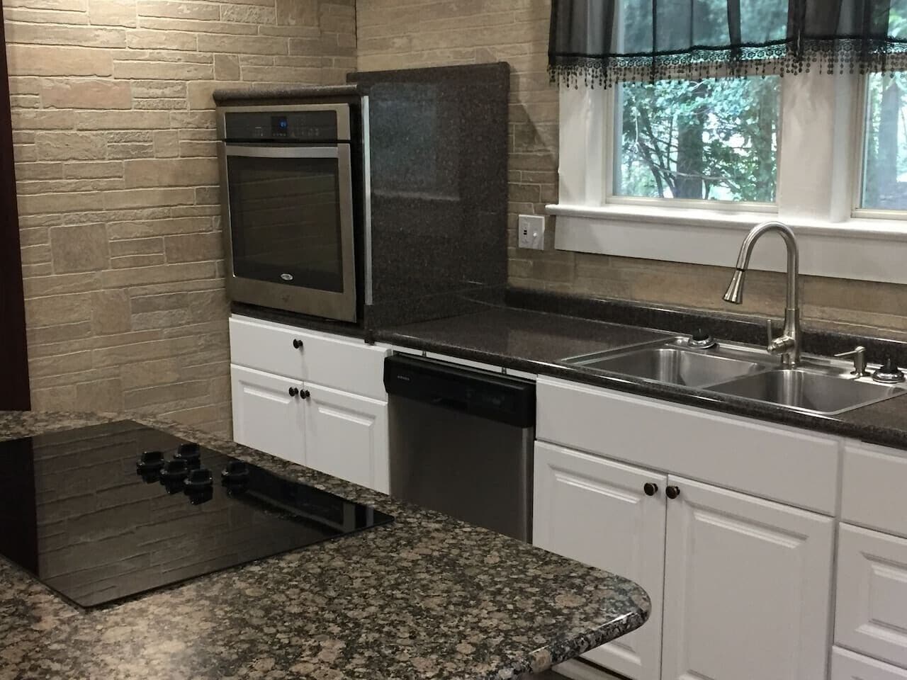 Una cocina con horno empotrado, lavavajillas, encimeras de granito, armarios blancos y una ventana con vistas a árboles.