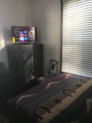 A room with a TV on a stand, a small chair, a bed with a patterned bedspread, and a window with blinds. - 3 bedroom central in Chattanooga (Chattanooga)