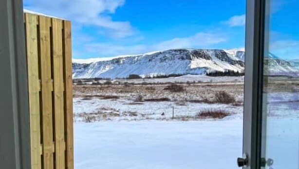 A view through a door with a wooden panel, leading to a snowy landscape with mountains in the distance. - Ferjukot Apartments (Sveitarfélagið Ölfus)