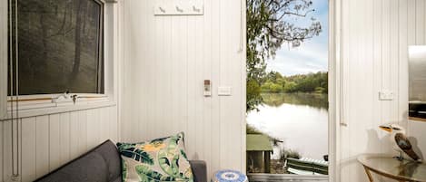 A room with a view of a lake, a sofa with a tropical pillow, a small table with a bird sculpture, and a window with a curtain.