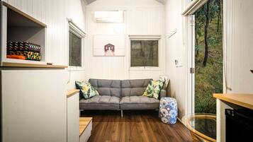 A compact room with a grey sofa, two windows, a small table, and a patterned stool.