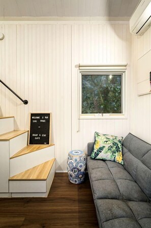 A room with a staircase, a grey sofa, a decorative vase, and a window with a view of trees. - Picturesque Lakefront Tiny House for Tranquil Glamping in Victoria (Flowerdale)