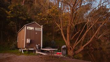 A wooden cabin with a bench and a table, surrounded by trees and a gravel path.
