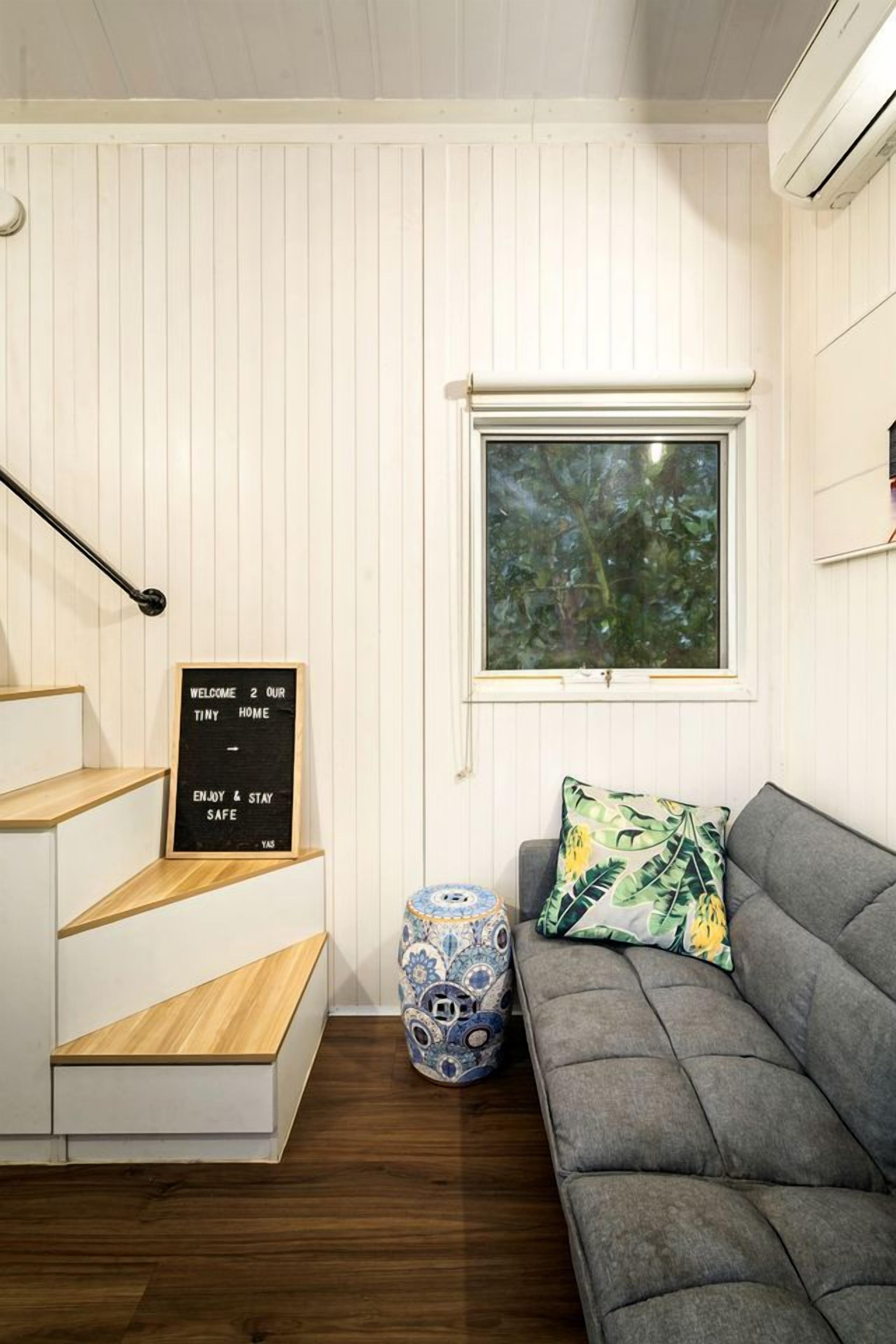 A room with a staircase, a grey sofa, a decorative vase, and a window with a view of trees.