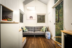 A compact room with a grey sofa, two windows, a small table, and a patterned stool.