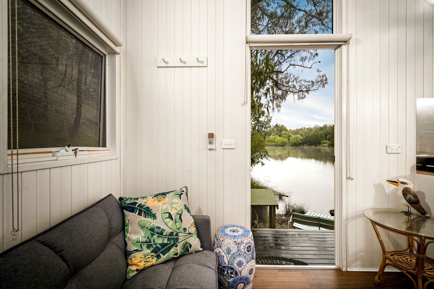 A room with a view of a lake, a sofa with a tropical pillow, a small table with a bird sculpture, and a window with a curtain.