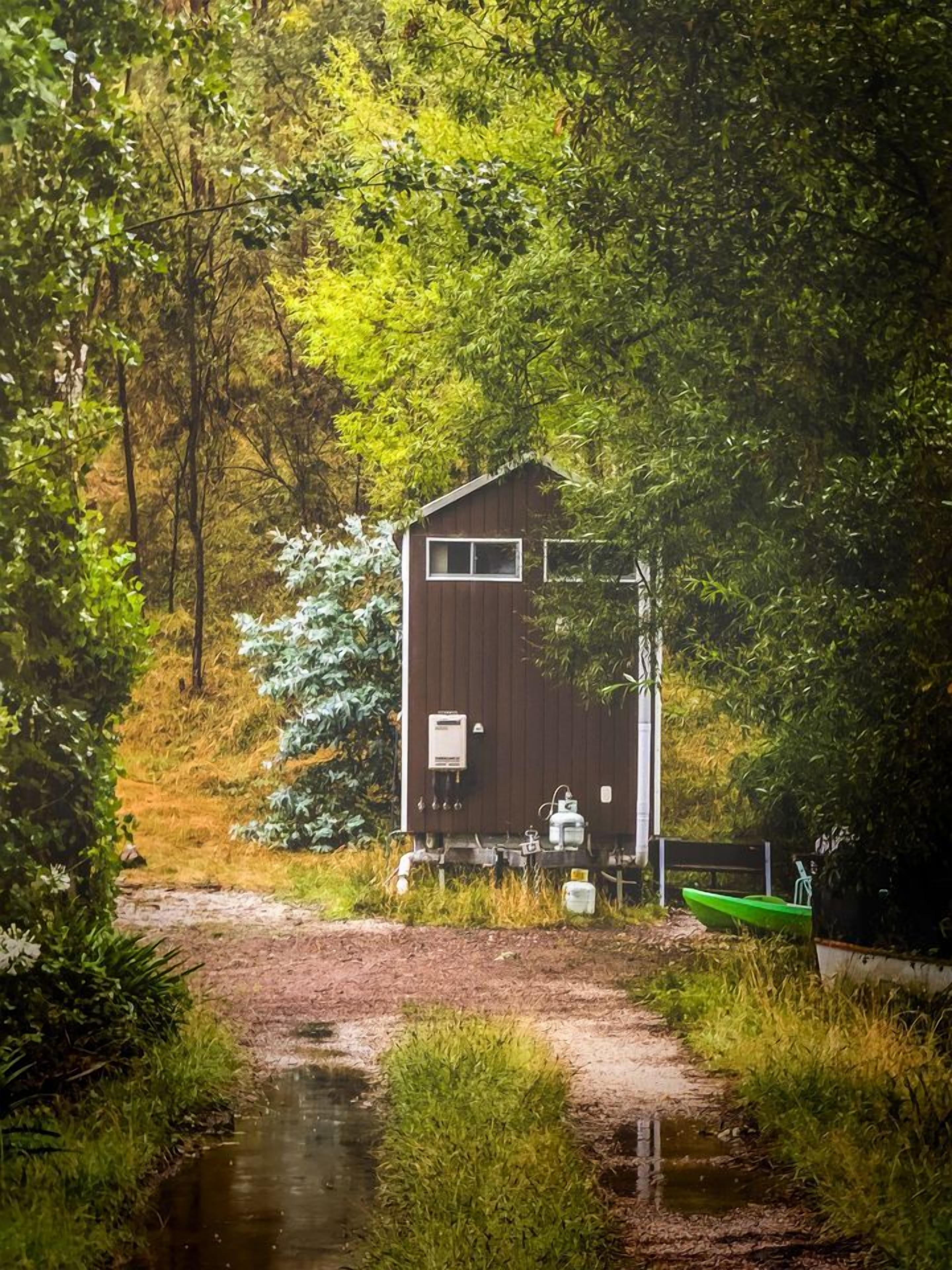 A wooden cabin with a small porch, surrounded by lush greenery and a stream.
