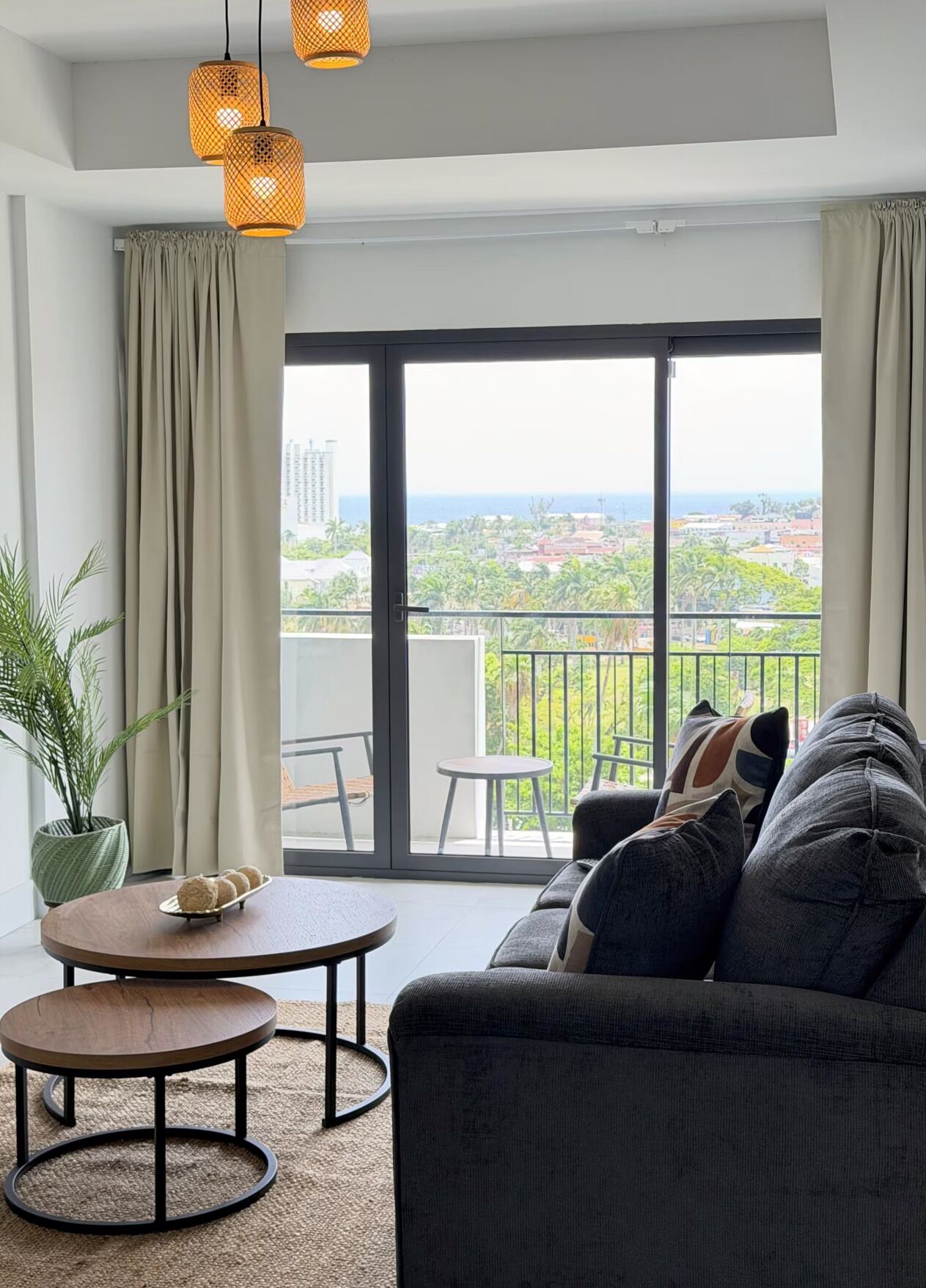 Uma sala de estar moderna com um sofá, uma mesa redonda pequena, uma planta em vaso e vista para uma varanda com guarda-corrimão.