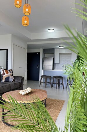 A modern living room with a sofa, a coffee table, and a kitchen area with a refrigerator and stools.