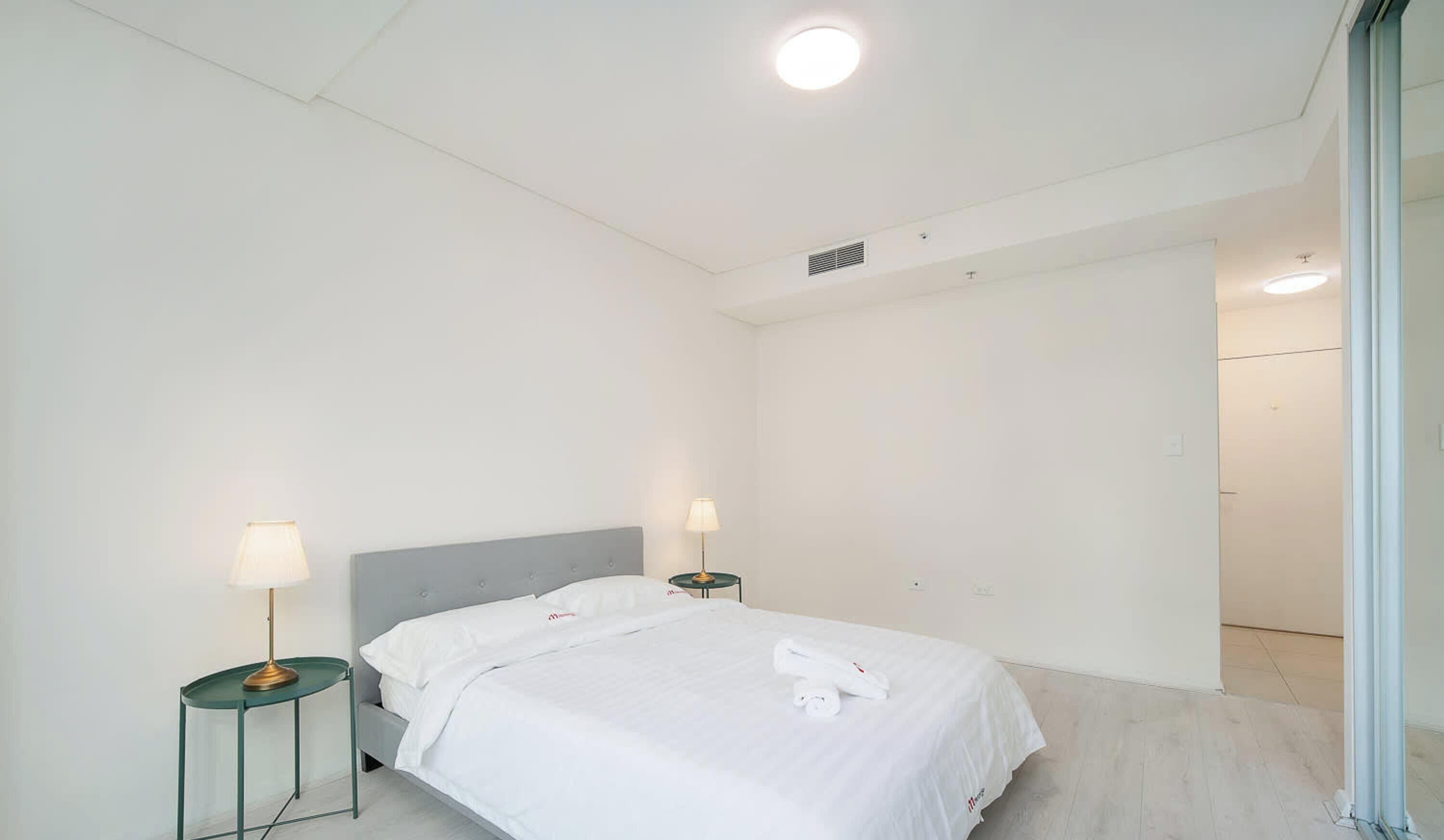 A hotel room with a bed, two bedside tables with lamps, a ceiling light, and a door leading to another room.