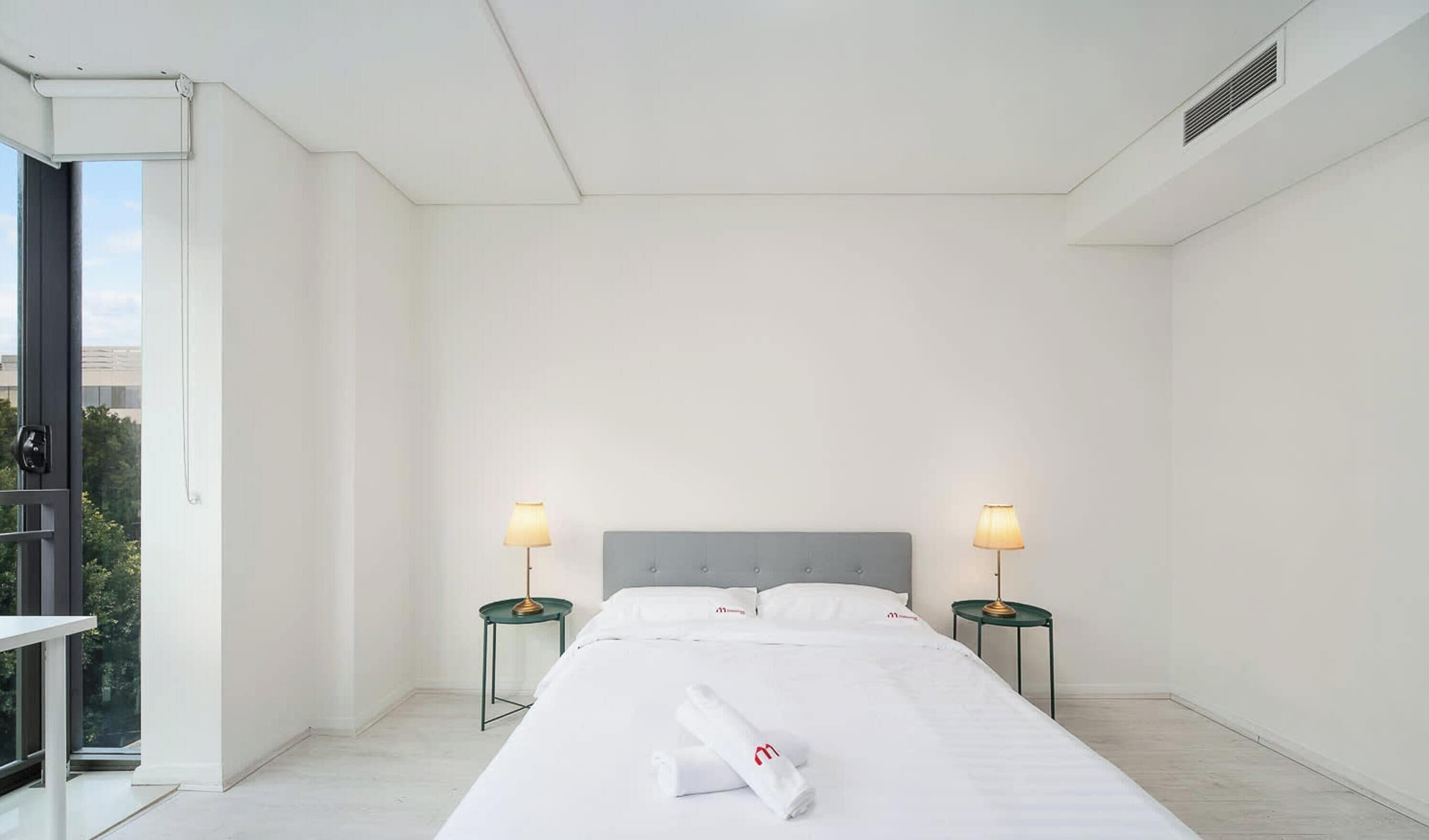 A hotel room with a large bed, two bedside tables with lamps, a window, and a view of greenery outside.