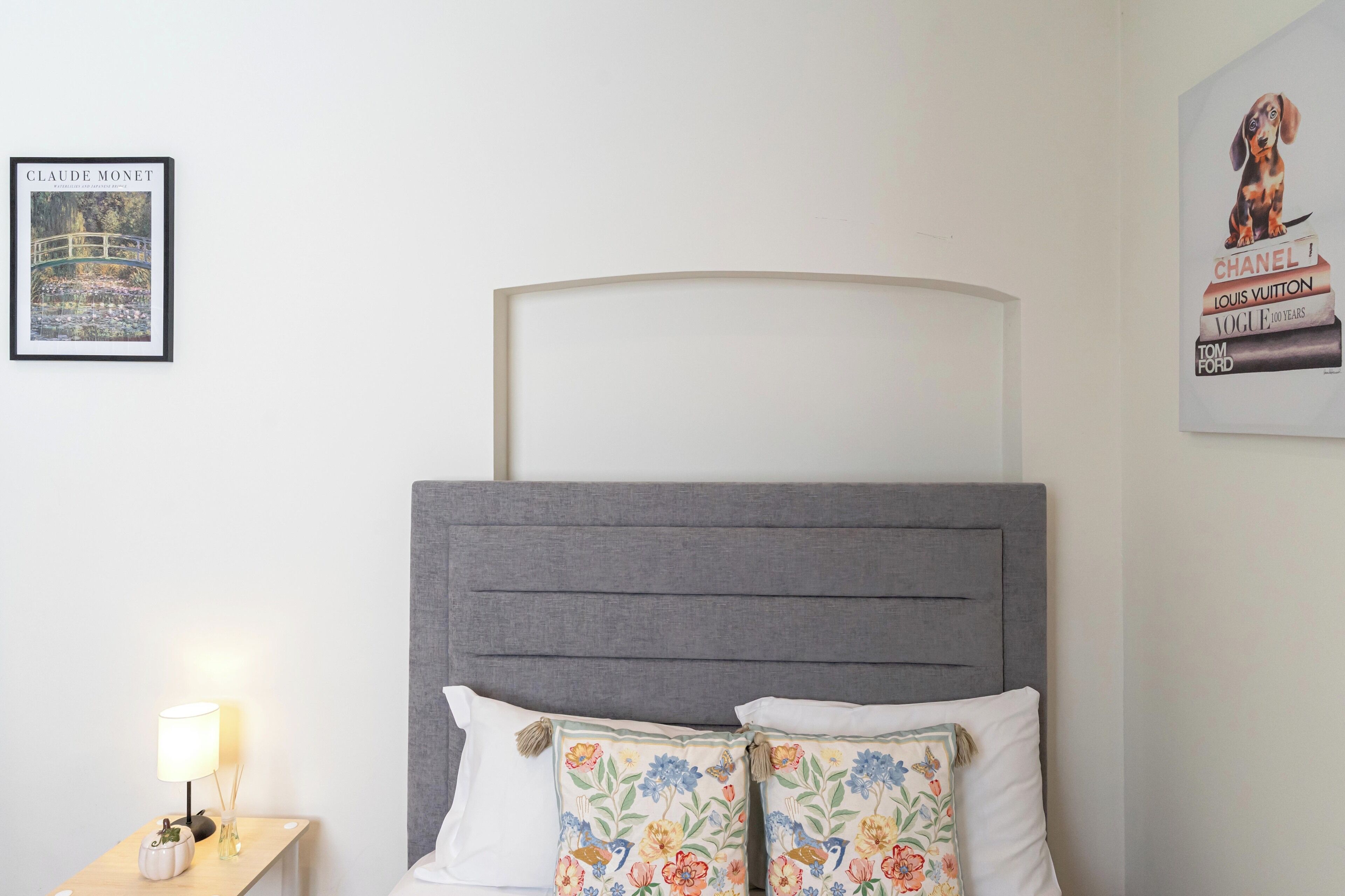 Ein Bett mit grauem Kopfteil, weißen Kissen und einem blumigen Kissen. Darüber hängt ein gerahmtes Kunstwerk, und auf dem Nachttisch steht eine Lampe.