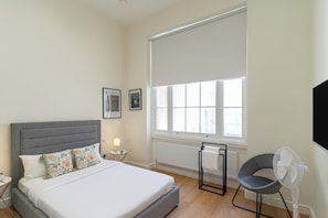Quarto com cama, uma cadeira, ventoinha, janela com persianas e televisão presa na parede.