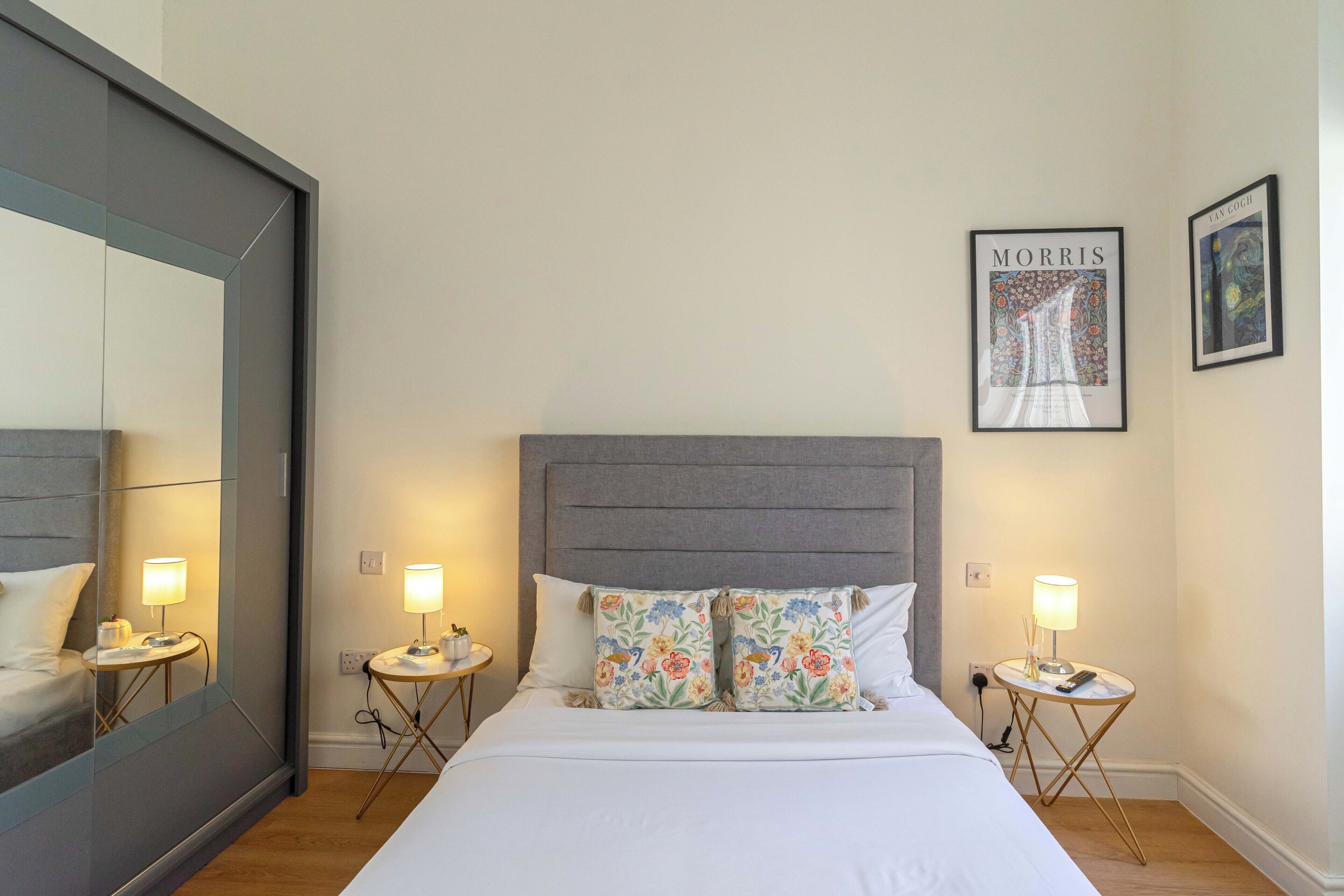 Une chambre d’hôtel avec un grand lit, deux tables de chevet avec des lampes, un grand miroir et deux œuvres d’art encadrées accrochées au mur.