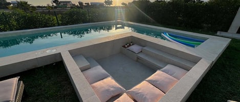 A modern outdoor pool area with a sunlit sky, a built-in seating area with cushions, and a view of the surrounding landscape.
