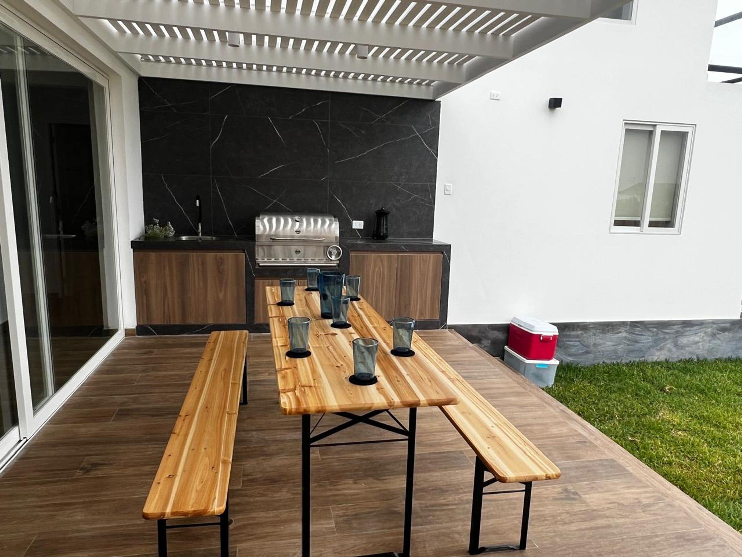An outdoor patio with a long wooden table and benches, a grill, and a modern kitchen area.