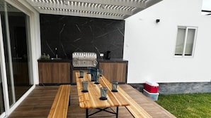 Un patio al aire libre con una larga mesa de madera y bancos, una parrilla y una cocina moderna.