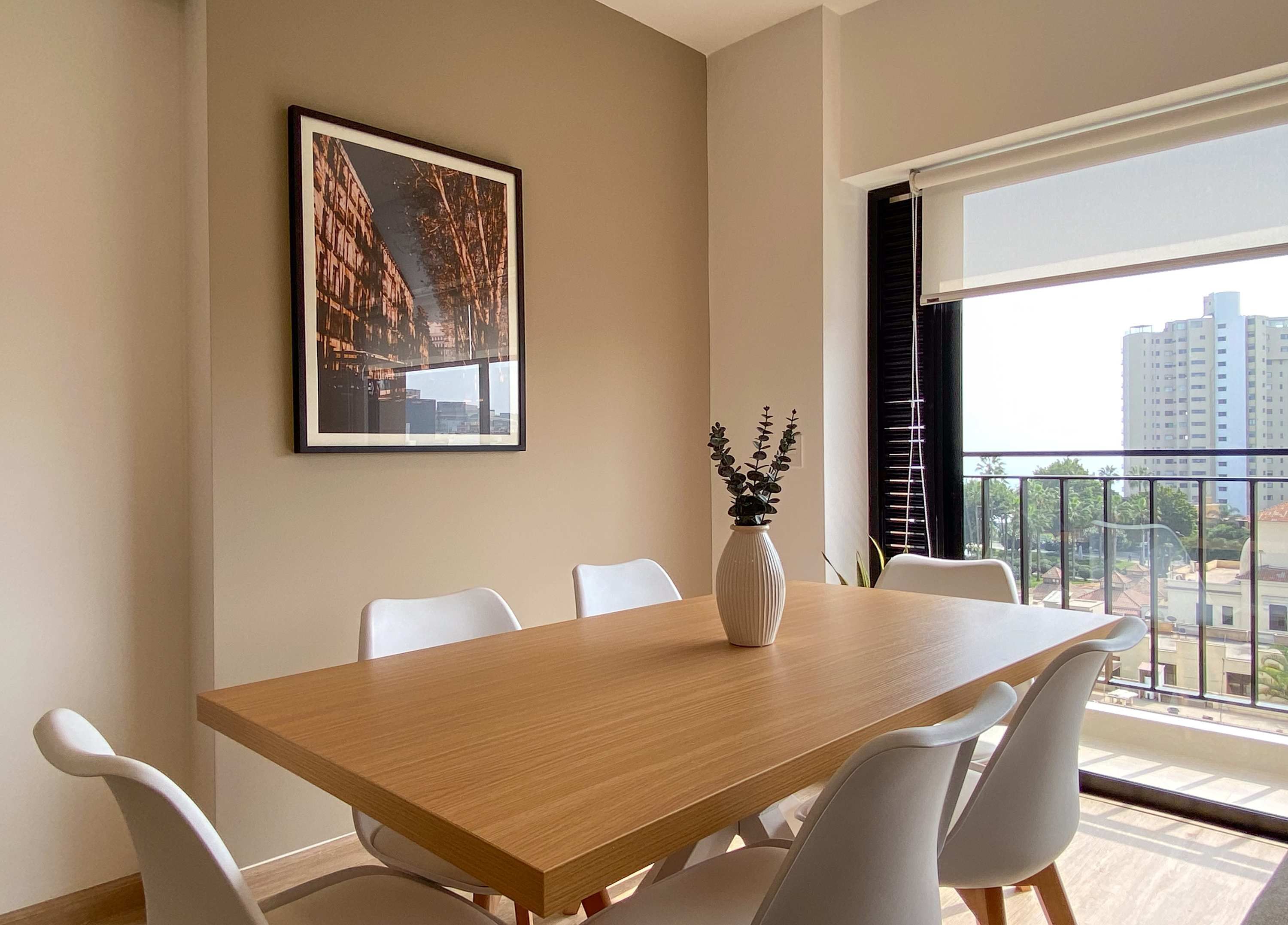 A modern dining area with a wooden table, white chairs, a vase with plants, a framed picture on the wall, and a balcony with a view of buildings.