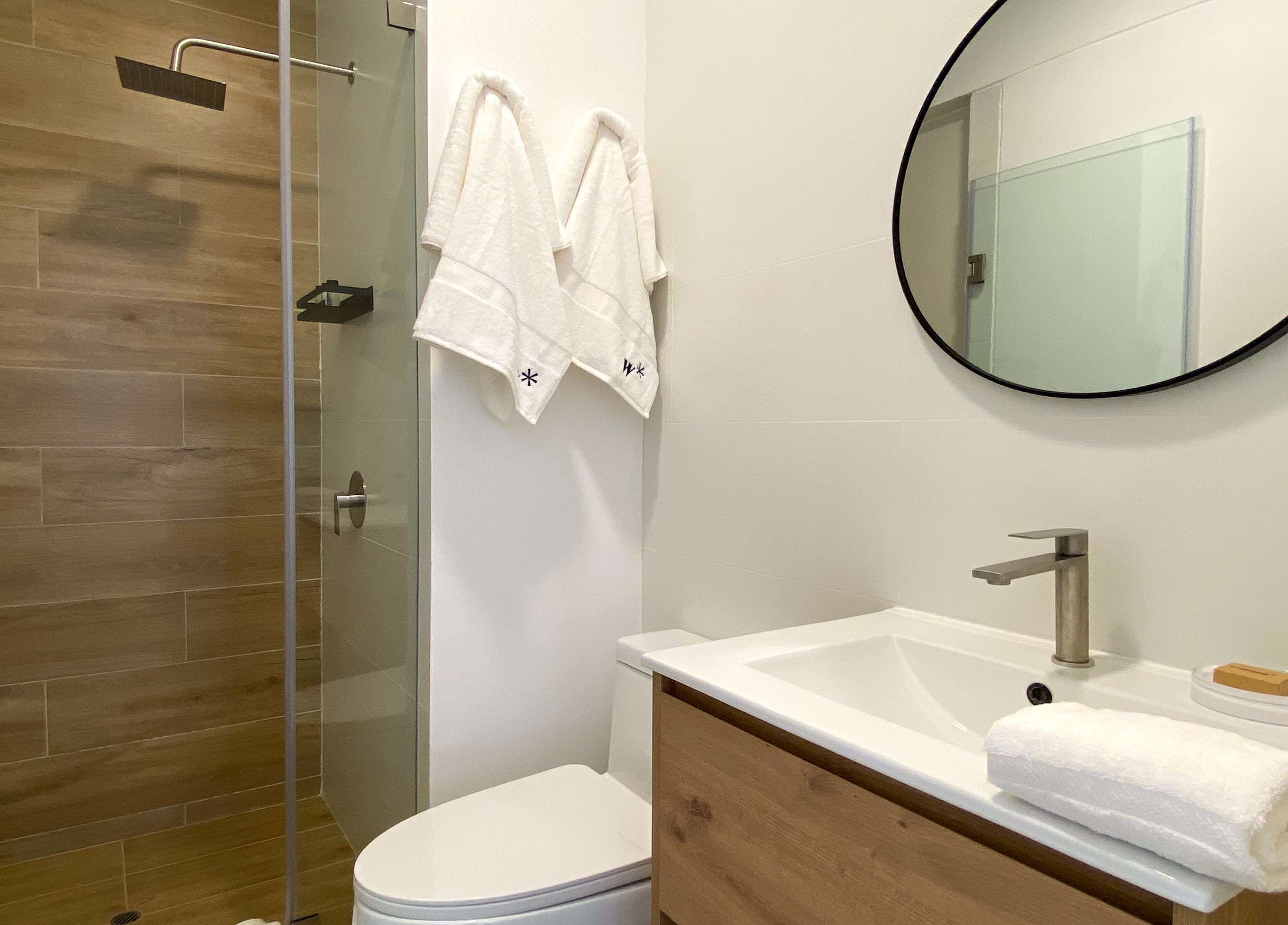 A modern bathroom with a glass shower enclosure, a white toilet, a wooden vanity with a white sink, and two white towels hanging on the wall.