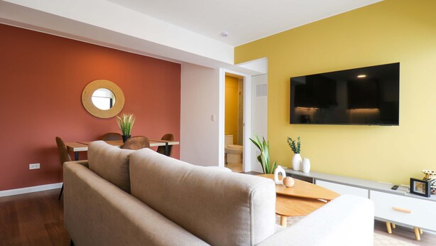 En moderne stue med sofa, rundt spejl, spisebord med stole og et fladskærms-tv på væggen.