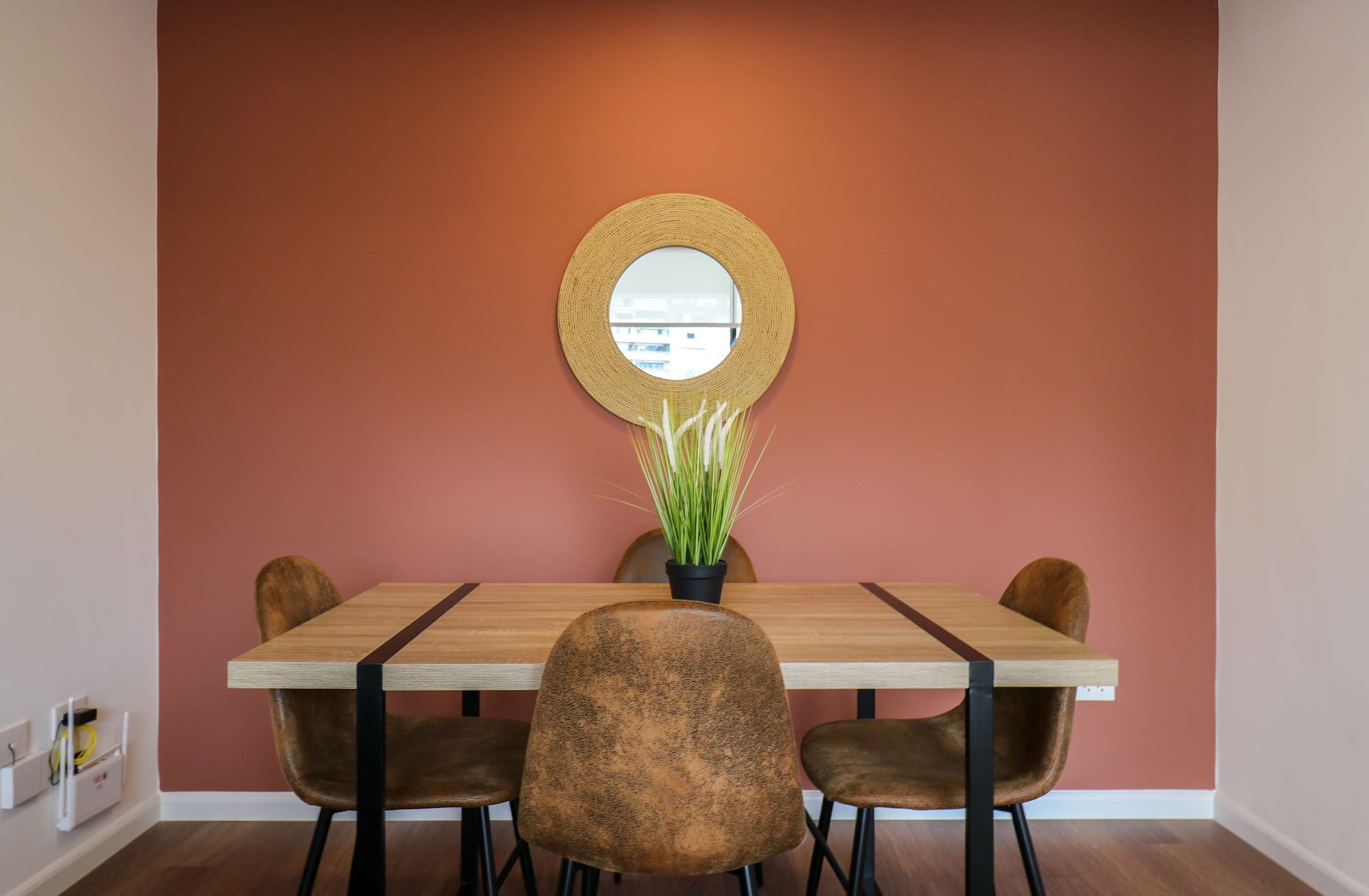 Een eetgedeelte met een houten tafel, vier stoelen, een ronde spiegel en een plant in pot.
