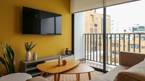 En moderne stue med gul væg, et ophængt fjernsyn, en sofa, et sofabord og en balkon med udsigt over byen.