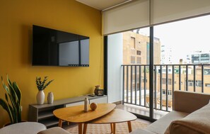 Ein modernes Wohnzimmer mit gelber Wand, einem an der Wand befestigten Fernseher, einer Couch, einem Couchtisch und einem Balkon mit Stadtblick.