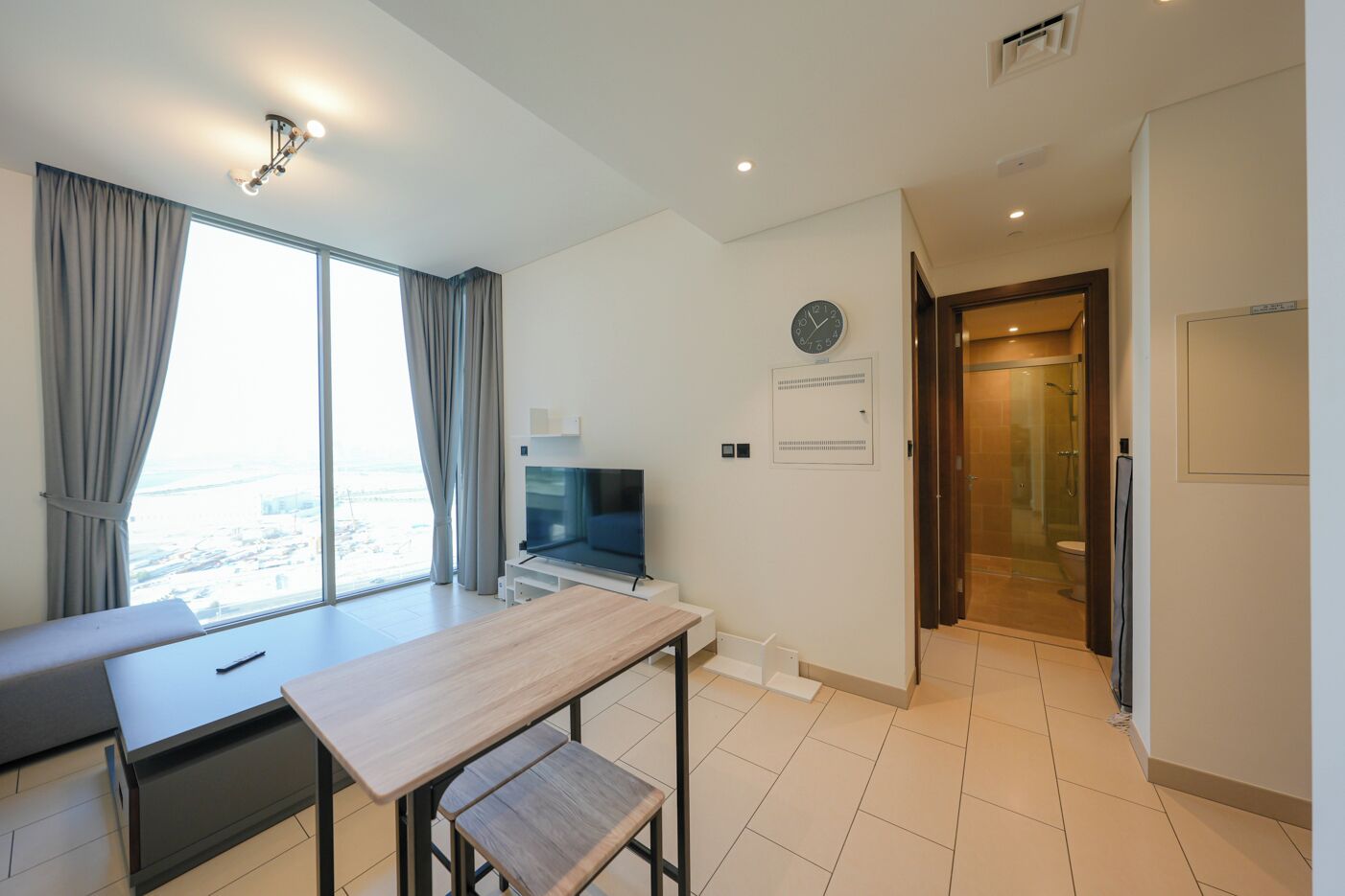 A modern hotel room with a dining area, a TV, a clock, and a shower area.