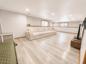 A spacious living room with a light-colored sofa, a green ottoman, a wooden side table, and a flat-screen TV on a wooden stand. - The Hartford - Spacious 4-bedroom house (Des Moines)