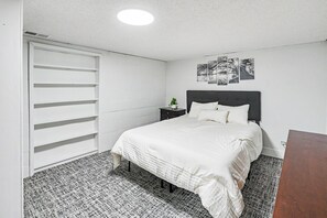 A bedroom with a bed, a nightstand, a wall-mounted shelf, and a ceiling light.