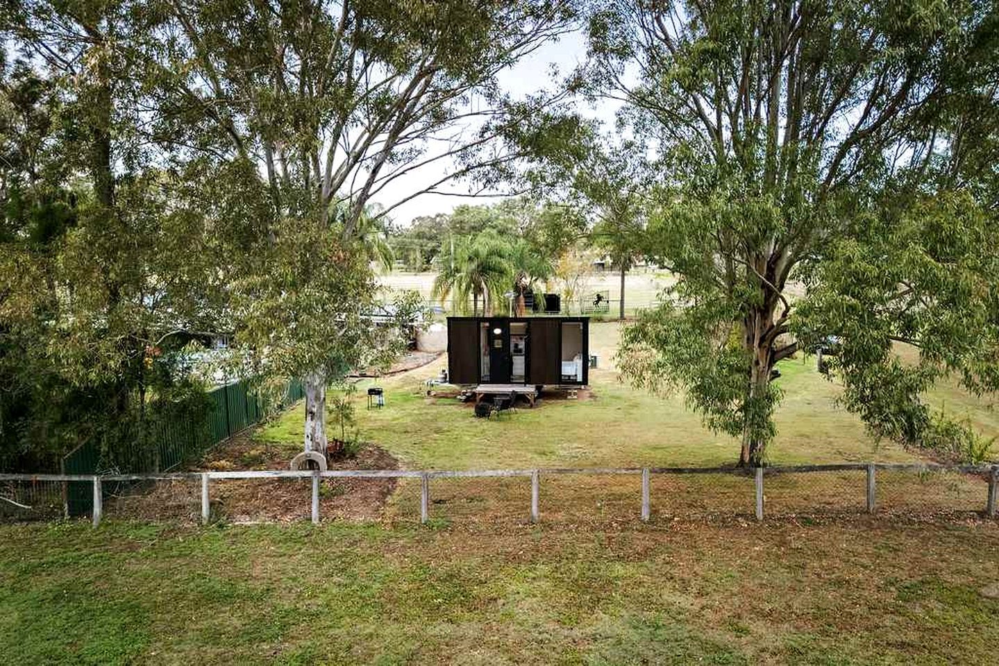 Een omheind terrein met een klein, zwart gebouwtje, mogelijk een tuinhuisje of opslagruimte, omringd door bomen.