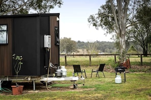 A small, dark-colored cabin with a metal structure, a water tank, and a chair outside.