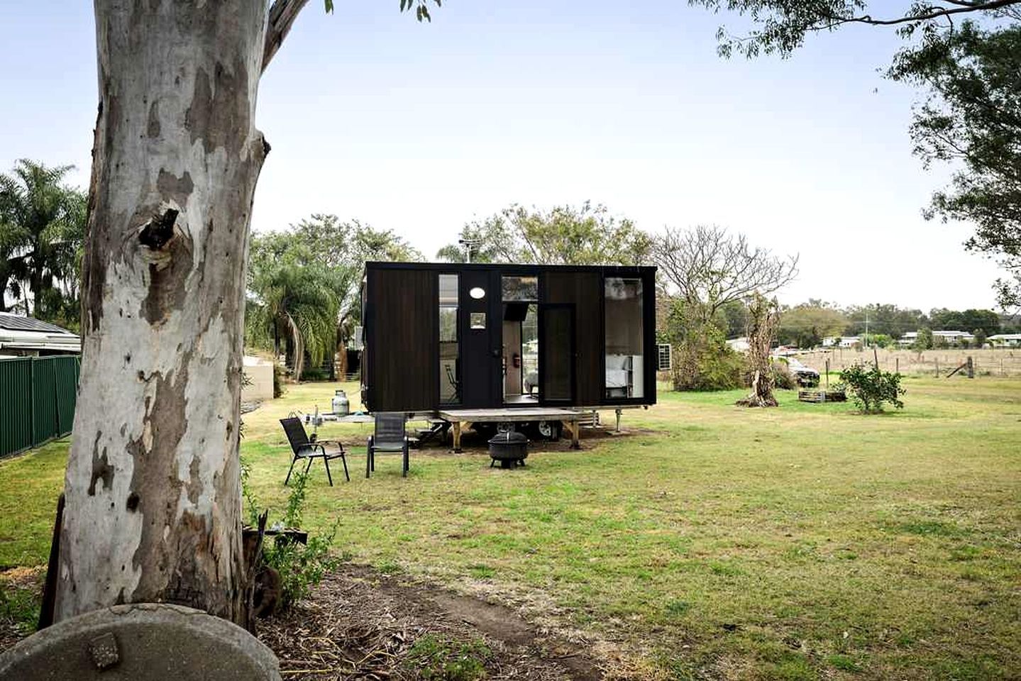 Een moderne, compacte stacaravan met een plat dak, gelegen in een gazon met een boom en een kleine tafel met stoelen.