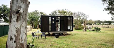 A modern, compact mobile home with a flat roof, situated in a grassy yard with a tree and a small table with chairs.