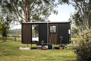 A small, dark-colored mobile home with a single visible window and a door, situated in a grassy area with trees and a fence in the background.