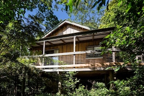 A two-story wooden cabin with a balcony, surrounded by dense forest.