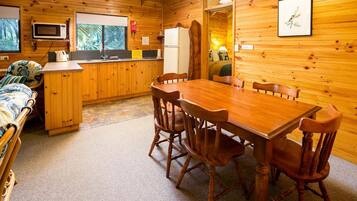 A wooden cabin interior with a dining table, chairs, a kitchen area, and a bed.